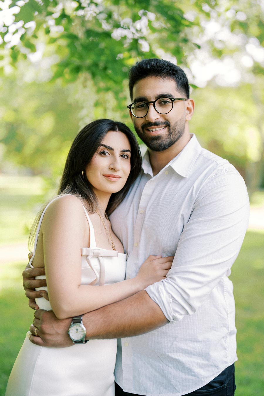 Couple embracing in a green park setting, both wearing white outfits and smiling, with lush trees in the background, evoking a sense of love and tranquility.