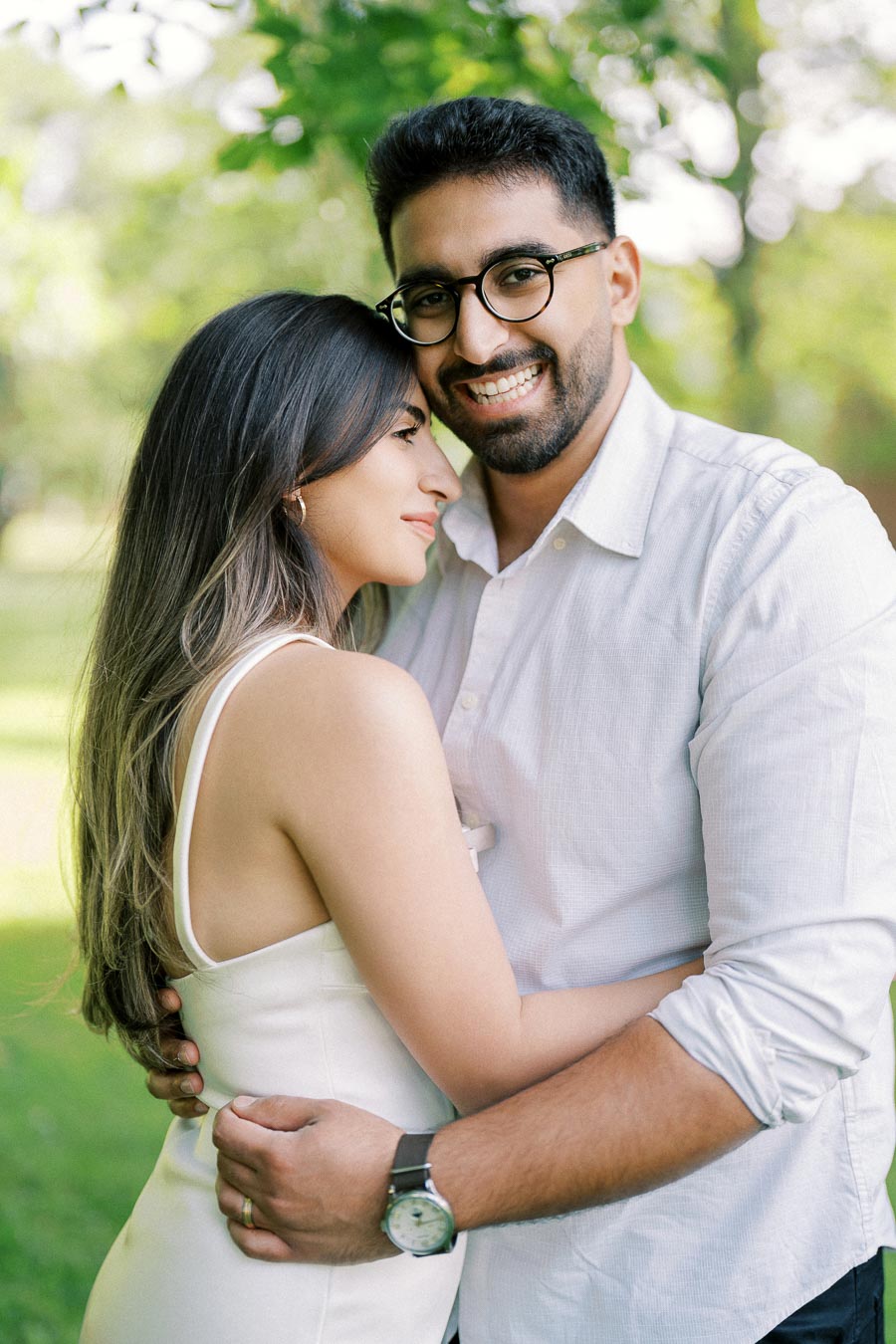 Couple embracing in a sunny park, wearing white outfits, surrounded by green trees and grass. The man has glasses and a watch, while the woman leans affectionately against him