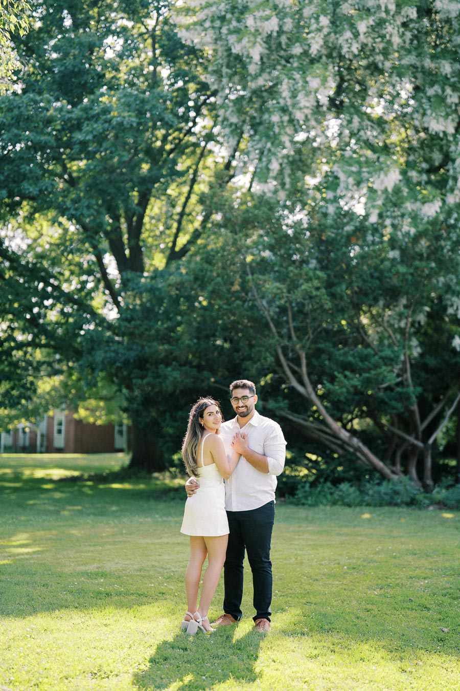 A couple embracing in a sunlit park with lush green trees in the background, both wearing white and looking content.