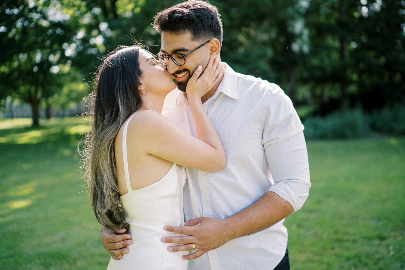 A couple embracing in a park setting with sunlight filtering through the trees, both smiling and dressed in light clothing, capturing a tender romantic moment.