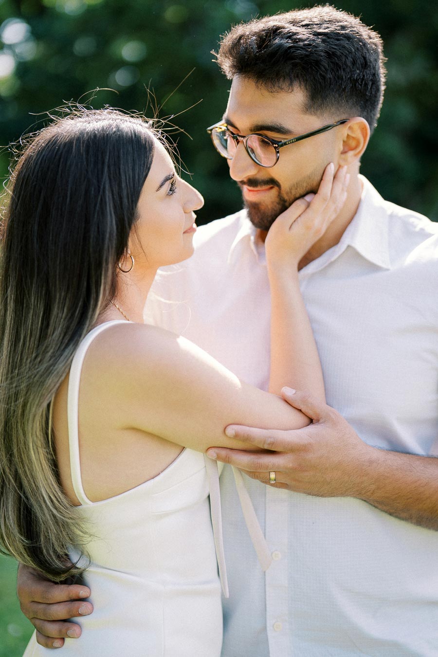 A couple gazing lovingly at each other outdoors, with soft sunlight filtering through the trees, both dressed in white, showcasing affection and intimacy.