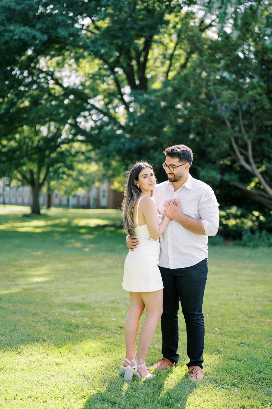 A couple standing closely on a sunny day in a lush green park, with the man smiling at the woman, who is wearing a white dress and heels.