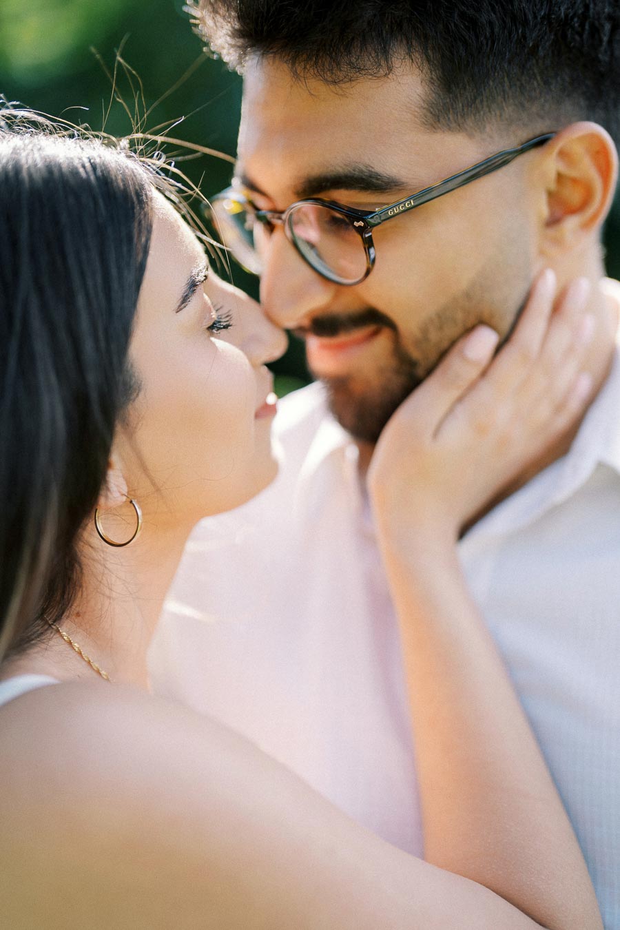A romantic close-up of a couple embracing outdoors, with the woman gently holding the man's face and both smiling lovingly at each other. The man is wearing glasses.
