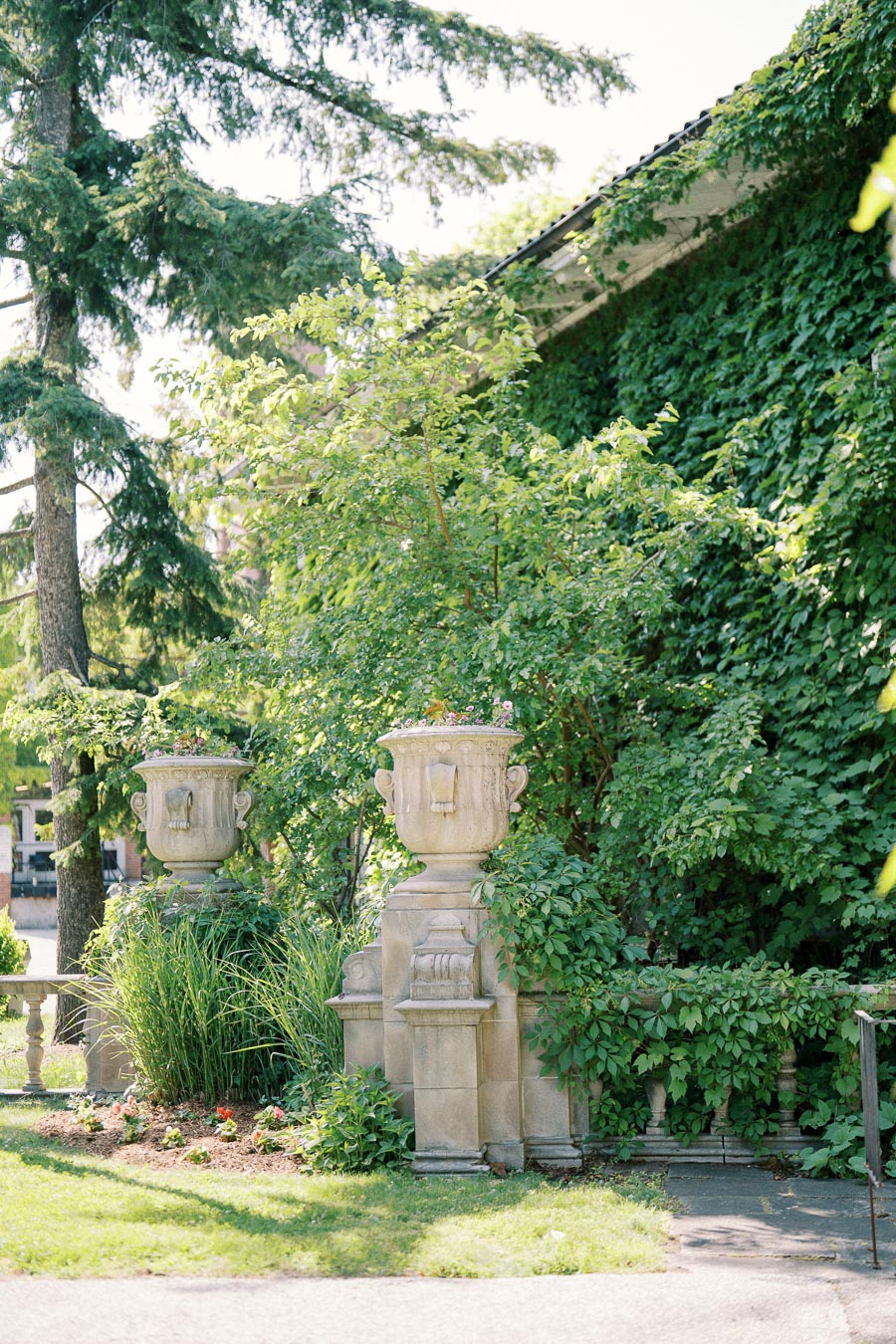 Decorative stone planters surrounded by lush greenery and ivy-covered walls in a garden setting, with sunlight filtering through the trees.