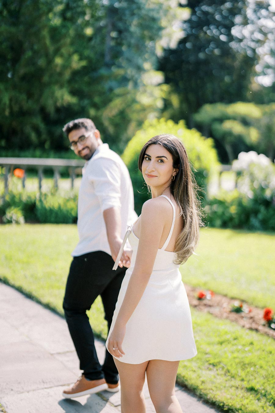 Young couple walking in a sunny garden, woman in a white dress looking back with a smile, lush greenery and bright flowers in the background.