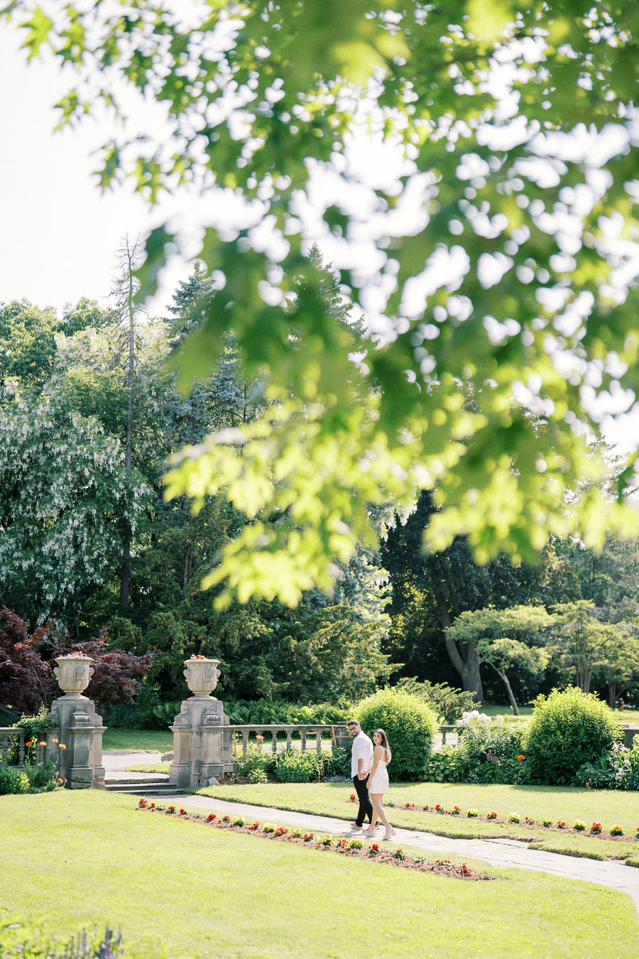 A couple walking hand in hand through a lush garden with vibrant greenery and stone urns in the background, under a canopy of sunlit leaves.