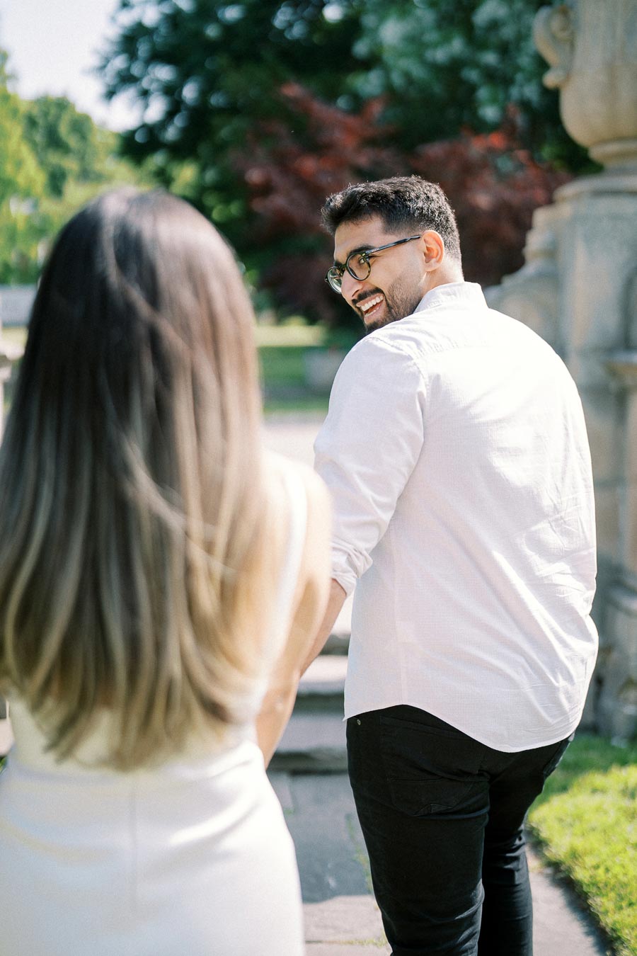 A man in a white shirt turns back, smiling at a woman with long hair outdoors. The sunlit scene is surrounded by green trees, suggesting a happy moment in a park setting.