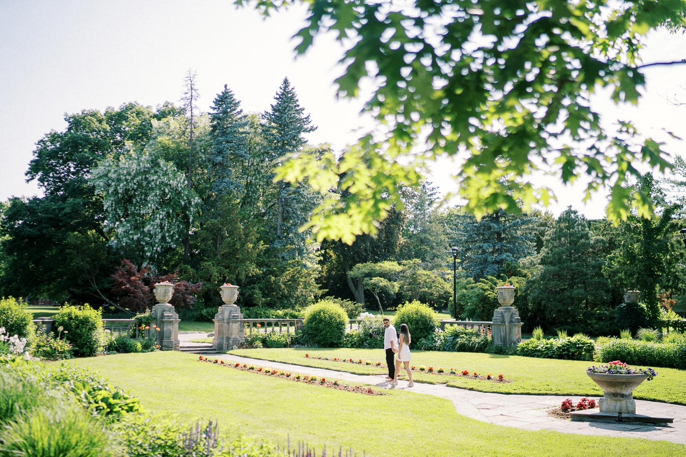 A couple walking hand in hand through a lush, green garden with stone urns and vibrant flowers, under the shade of large trees on a sunny day.