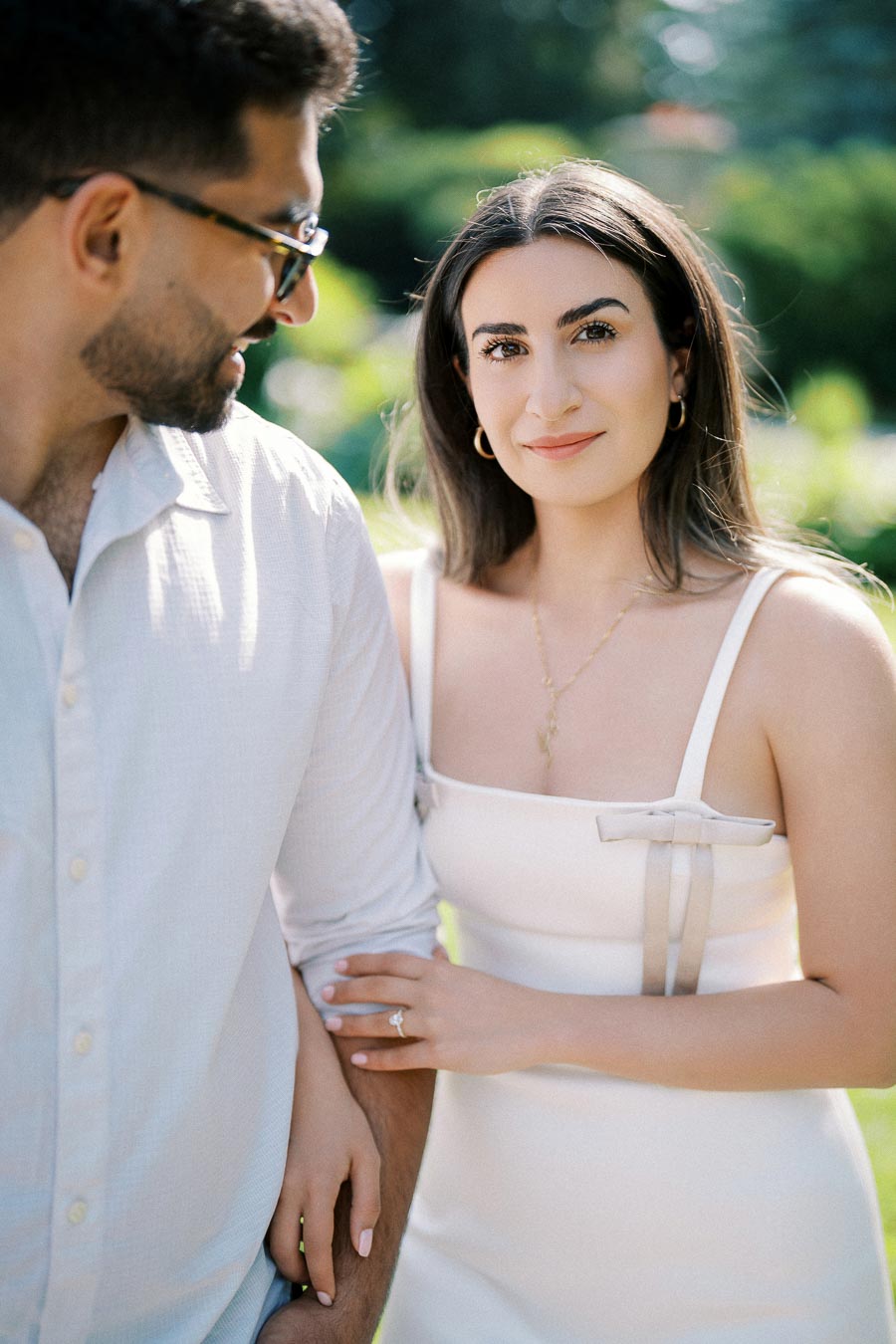A couple standing outdoors with the woman smiling at the camera while holding the man's arm, wearing a white dress and a ring, surrounded by a lush green background.