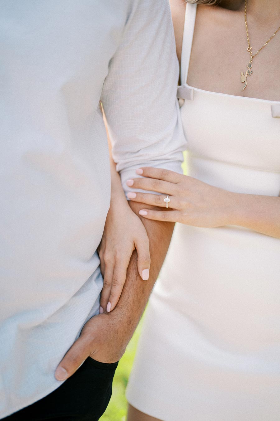 A close-up image of a couple embracing, with the woman wearing a diamond engagement ring on her ring finger. She is gently holding the man’s arm, both dressed in light-colored clothing, symbolizing love and commitment.