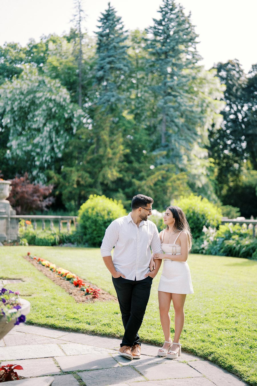 A couple walking in a lush garden, holding hands and smiling at each other, with vibrant greenery and flower beds in the background on a sunny day.
