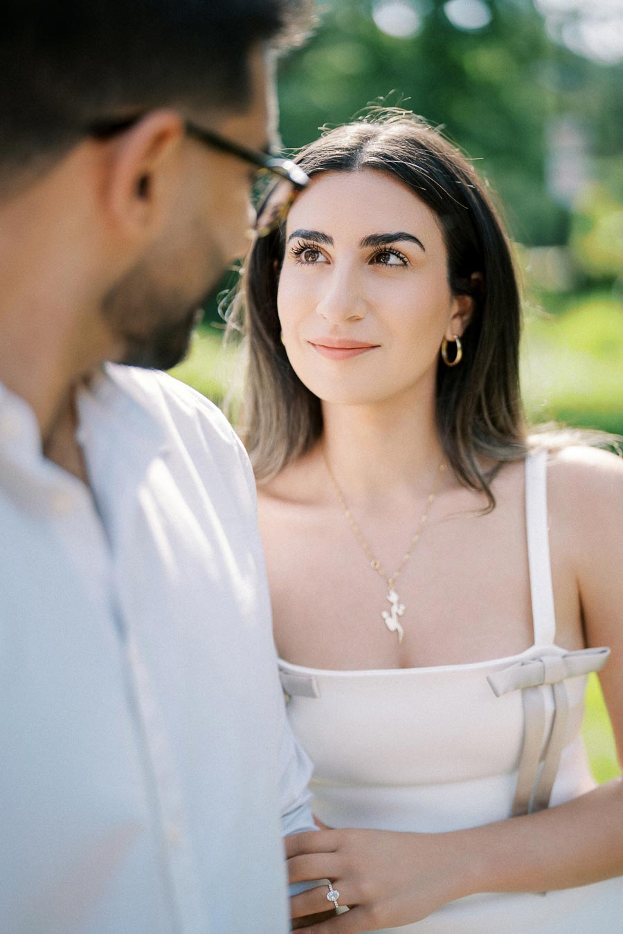 A woman in a white dress with hoop earrings and a necklace looks at a man in glasses, with a green, blurred outdoor background.