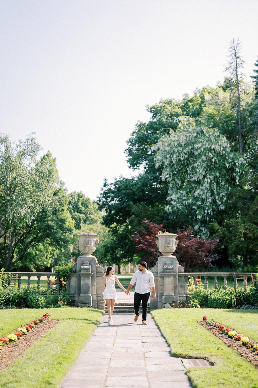 A couple walks hand in hand down a stone pathway flanked by colorful flowerbeds and ornate stone urns in a serene garden setting with lush greenery and clear blue sky.