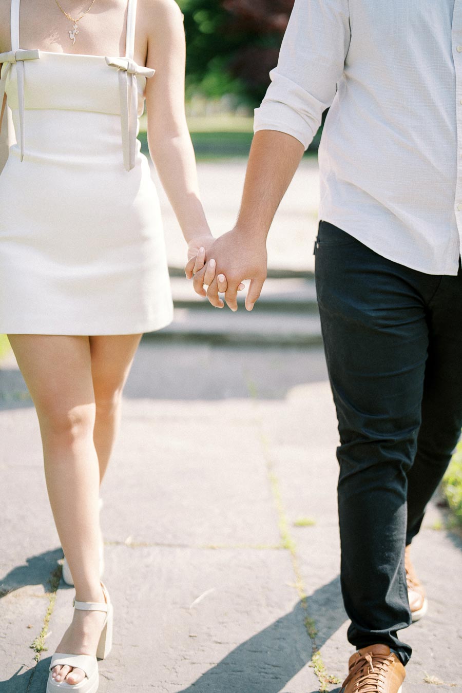A couple holding hands while walking outdoors, with one wearing a white dress and the other in dark trousers and light shirt, symbolizing romance and connection on a sunny day.