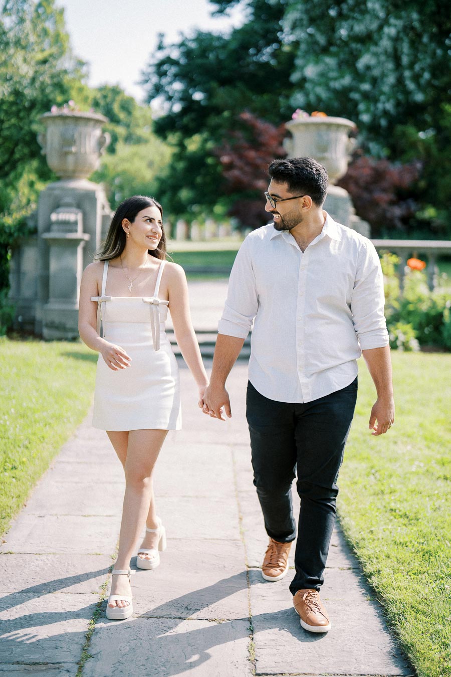 A couple holding hands and walking along a sunny garden path, surrounded by lush greenery and decorative stone urns.