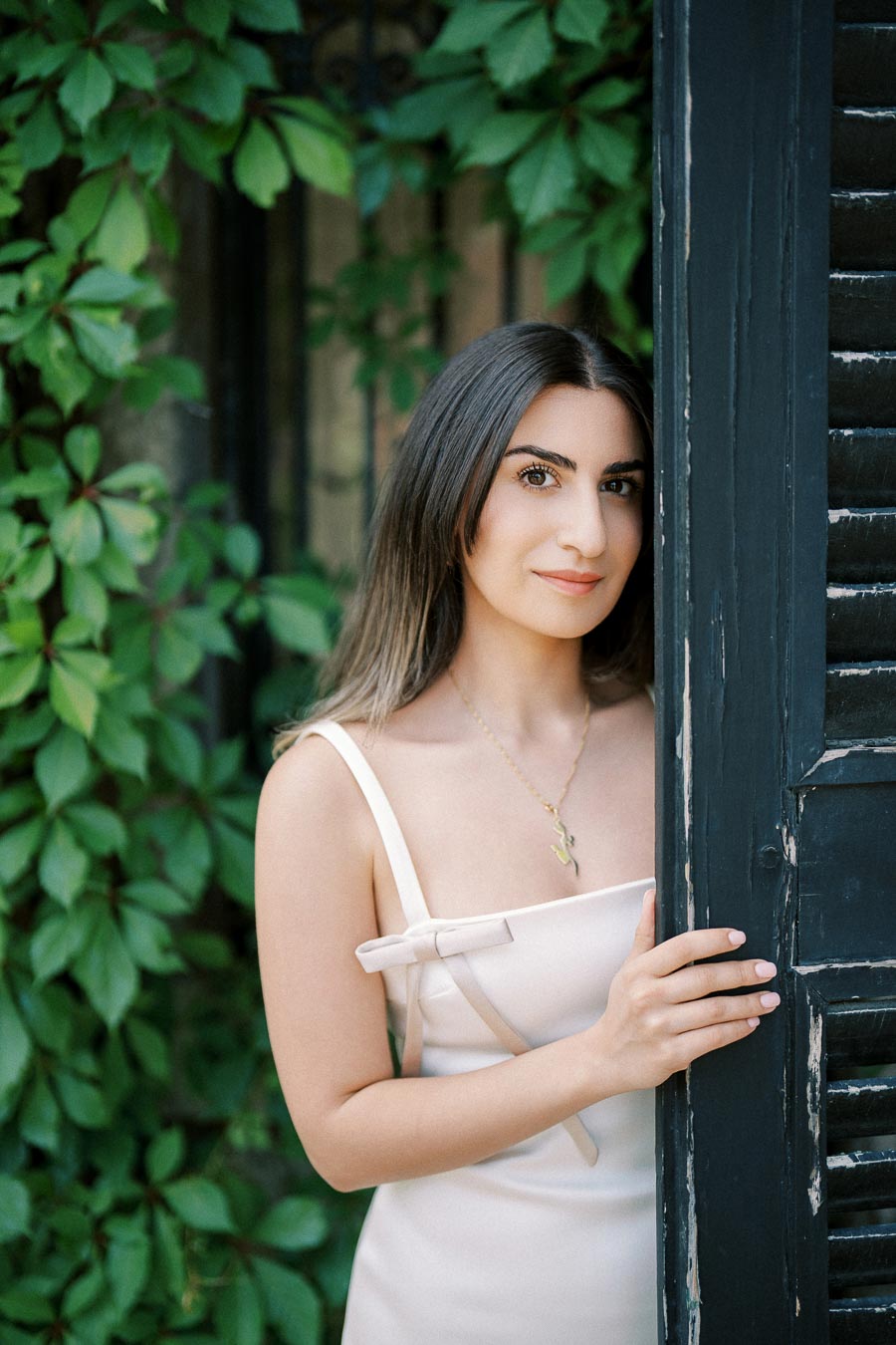 Young woman in a light-colored dress standing beside a rustic black door framed by lush green leaves, creating a serene and natural setting.
