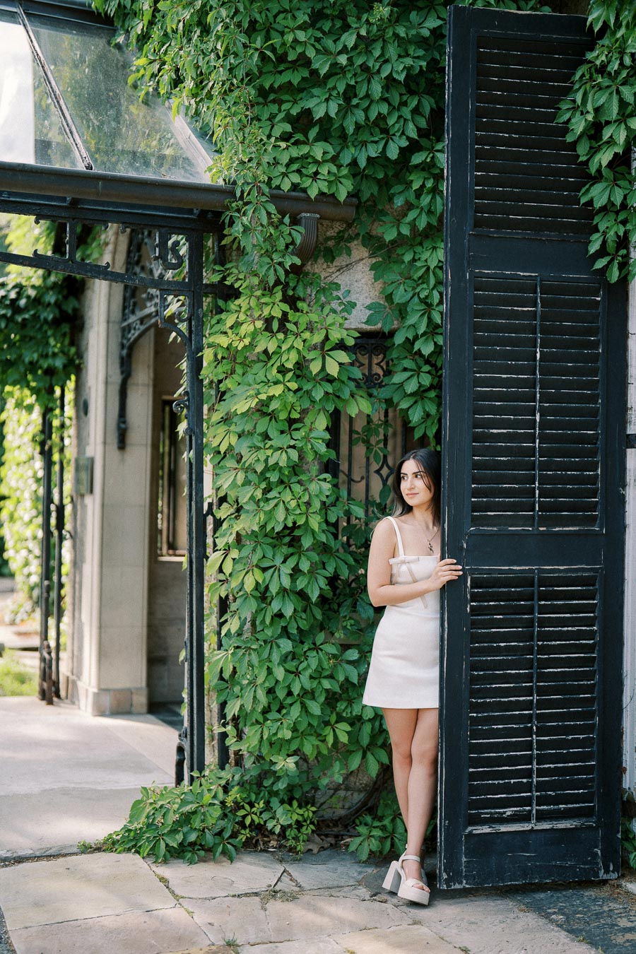 Young woman in a white dress standing by a large, rustic black shutter door with lush green ivy on a sunny day.