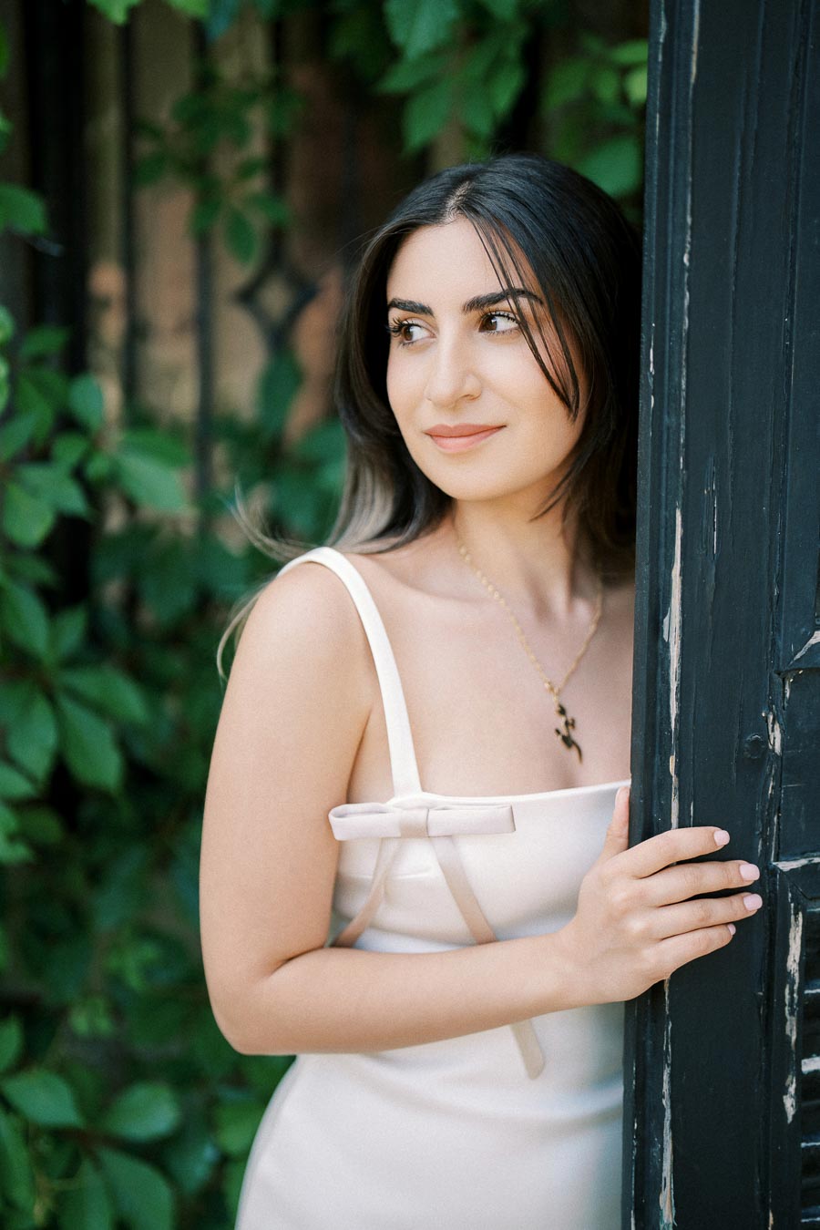 Young woman in a white dress smiling while standing by a rustic black door with green foliage background.