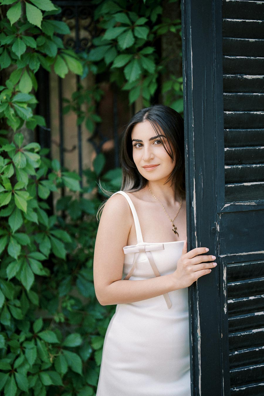 Woman in a white dress posing by a black wooden shutter with lush green ivy in the background.