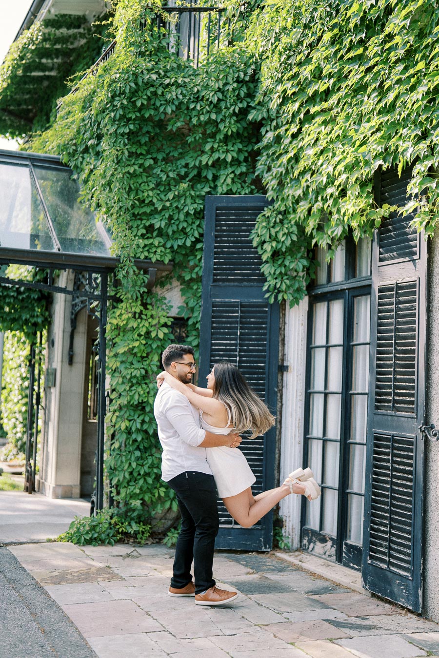 A couple embracing lovingly in front of an ivy-covered building with dark shutters, exuding a romantic and joyful atmosphere.