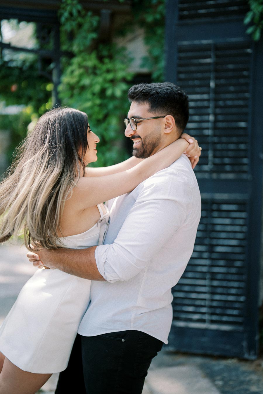 A happy couple embracing each other outdoors, surrounded by lush greenery, with the woman in a white dress and the man in a white shirt, both smiling warmly at each other.