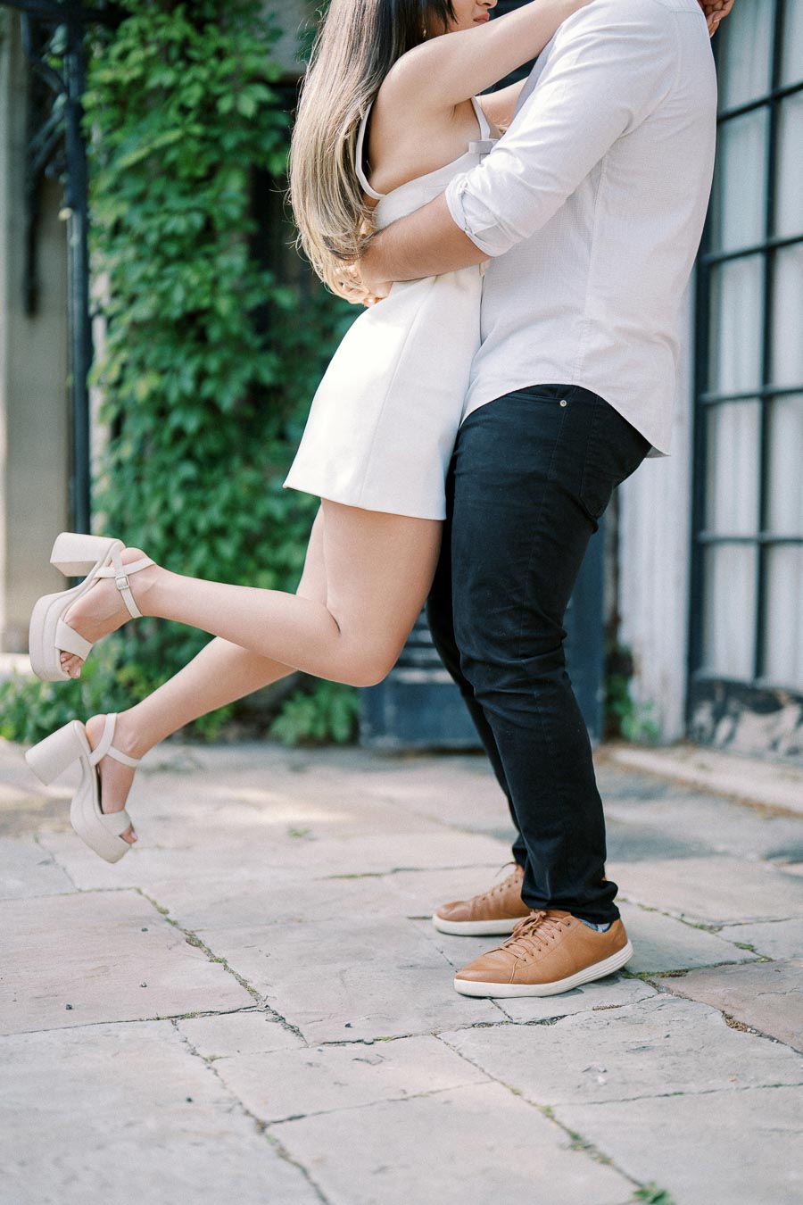 A couple embraces romantically outdoors, with the woman in a white dress and platform heels lifting her leg as the man holds her closely, set against a backdrop of green foliage and large windows.