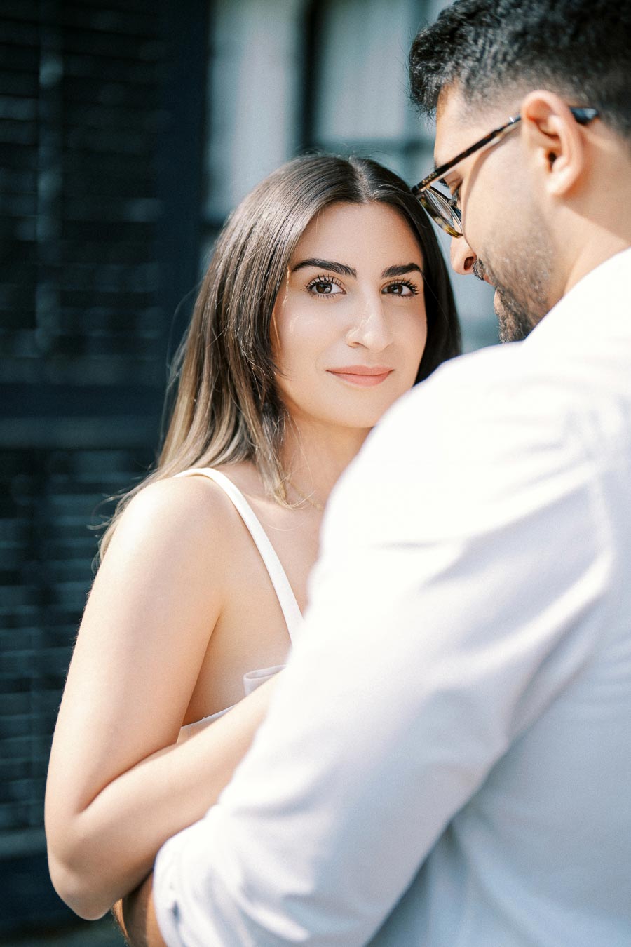 A couple engaged in a tender moment, with a woman with long brown hair looking at the camera and a man in glasses standing close to her in a softly lit outdoor setting.
