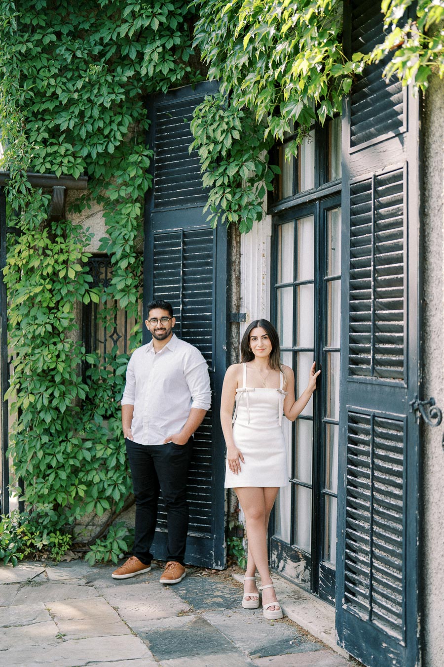 A couple posing fashionably in front of a historic building with black wooden shutters and lush green ivy, capturing a serene outdoor moment.