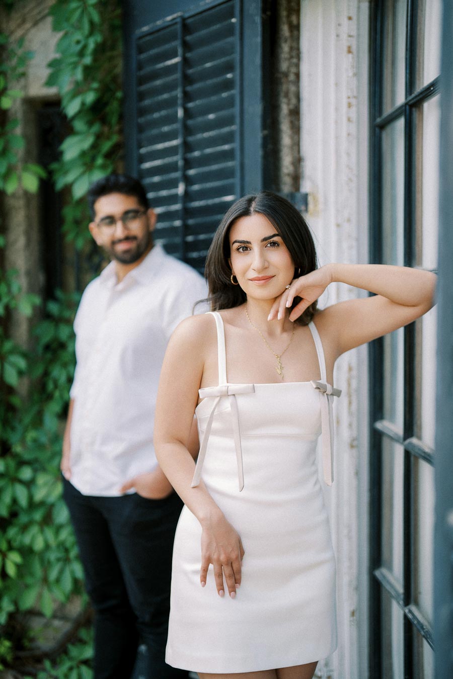 Young woman in white dress posing near a window with a man in the background, surrounded by greenery.