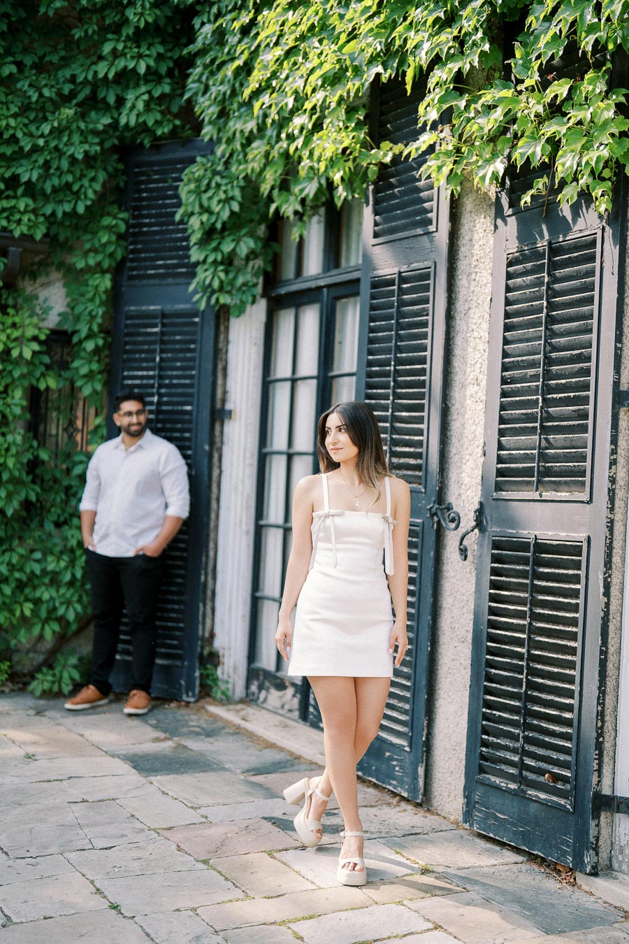 A stylish woman in a white dress walks in front of a vine-covered wall with black shutters, while a man in a white shirt stands in the background, creating a romantic, outdoor setting.