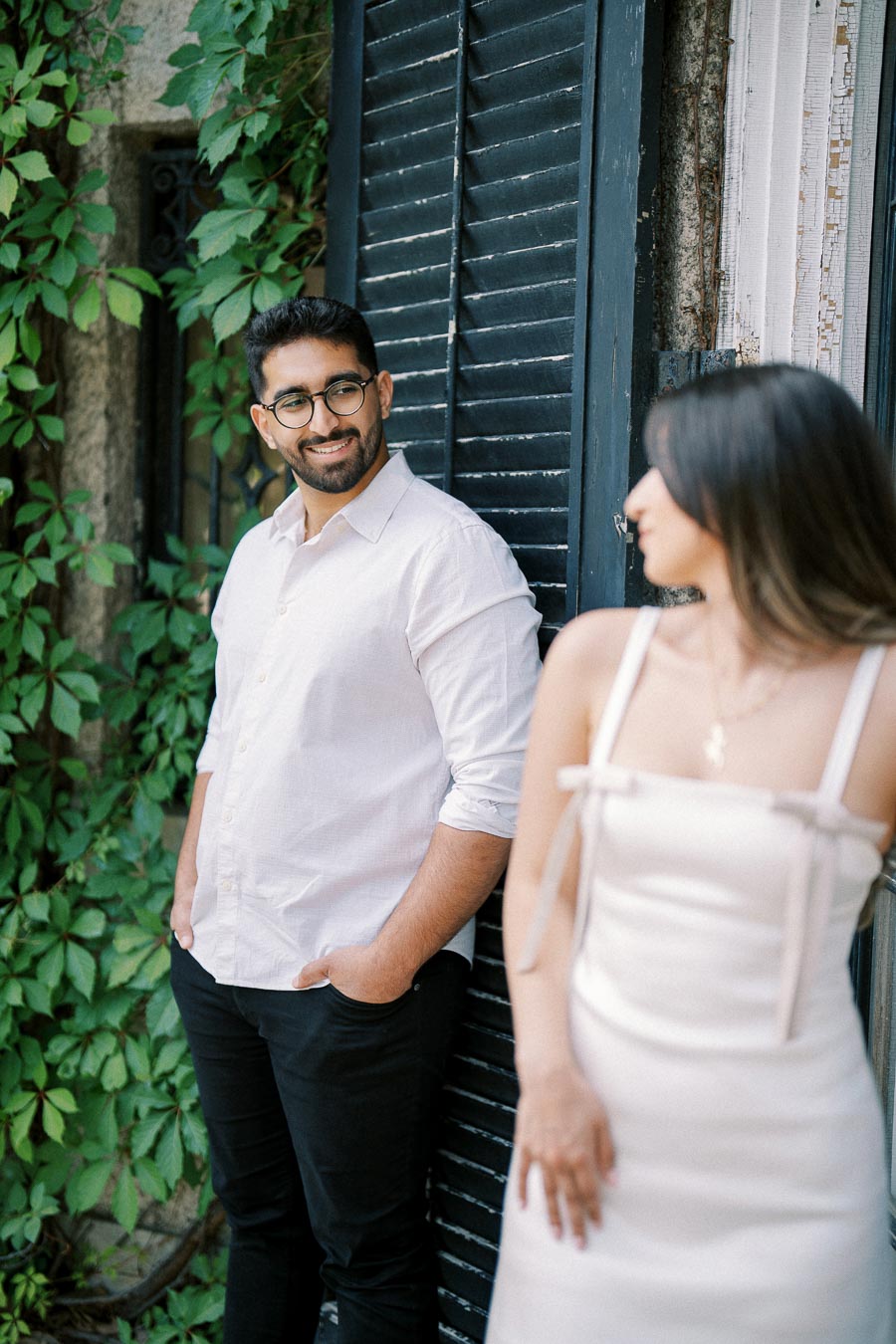 A man in a white shirt smiles at a woman in a white dress. They stand outdoors near a wall covered in green vines and a black shutter, creating a romantic and natural scene.
