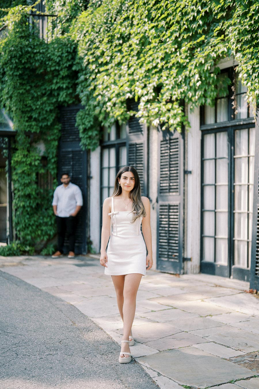 Young woman in a white dress walking confidently on a stone path with ivy-covered building in the background.