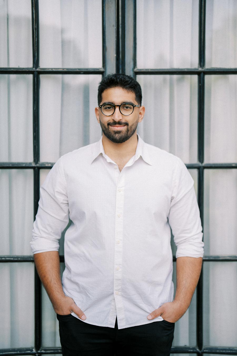 A man with a beard and glasses stands confidently in front of a large window, wearing a white shirt and dark pants.