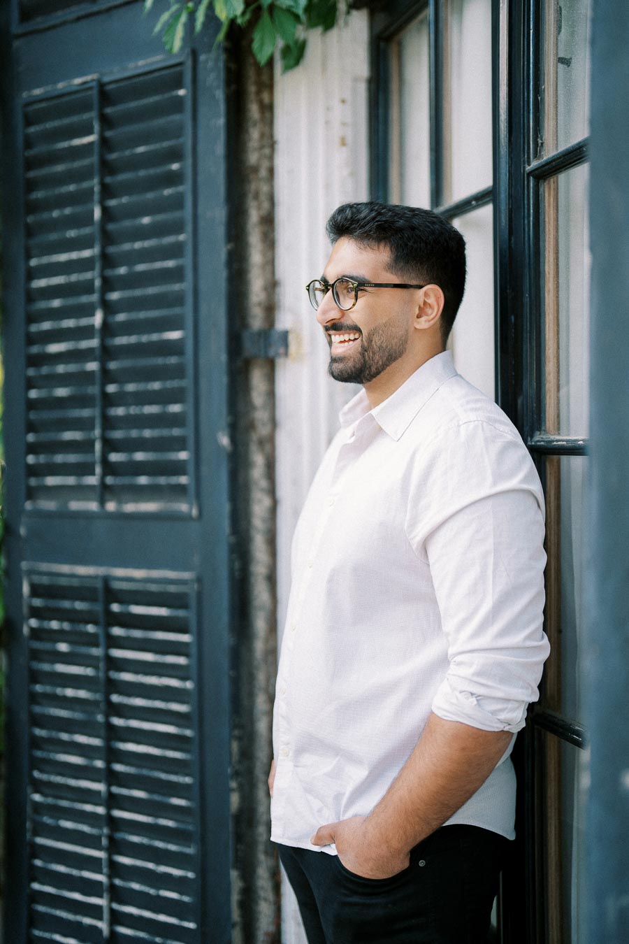 A man in a white shirt smiling while leaning against a rustic blue shuttered window, outdoors.