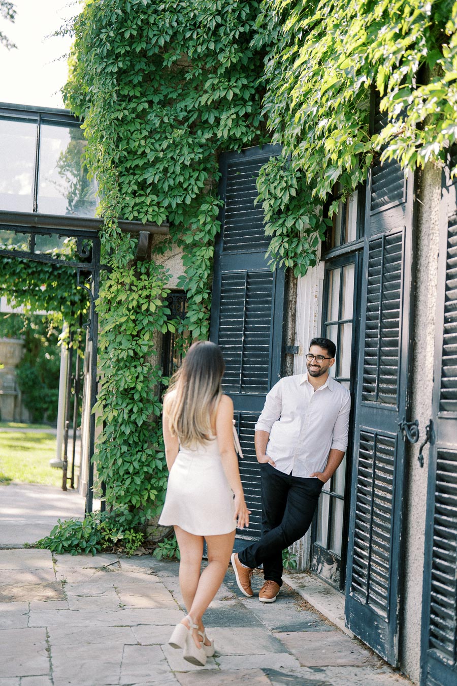 Man and woman interacting outdoors near a rustic building with black shutters, surrounded by lush green ivy.