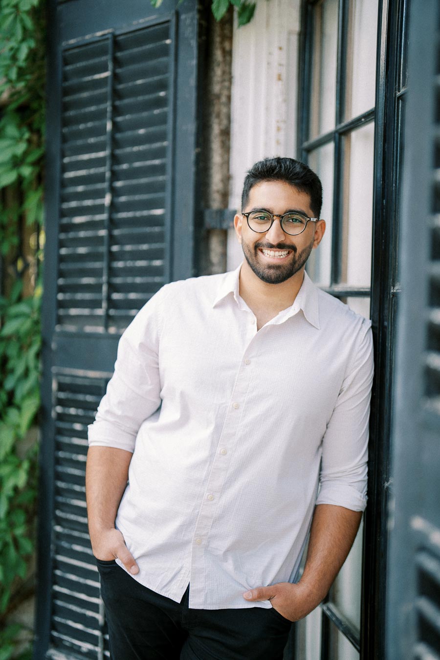 Smiling man wearing glasses and a white shirt, leaning against a wall with black shutters and greenery in the background.