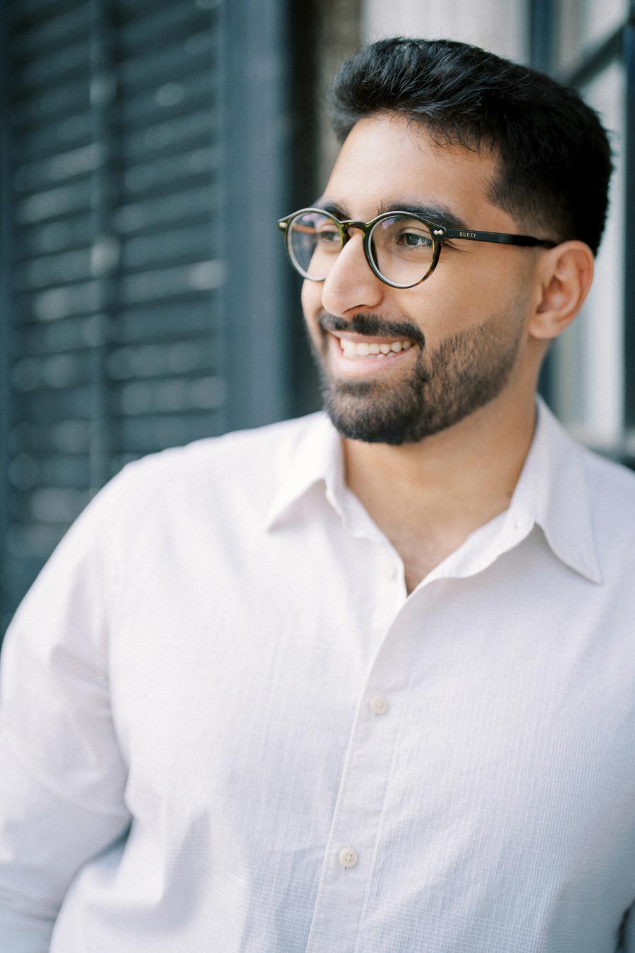 A man with glasses smiling and looking to the side, wearing a white button-up shirt, in a bright outdoor setting.