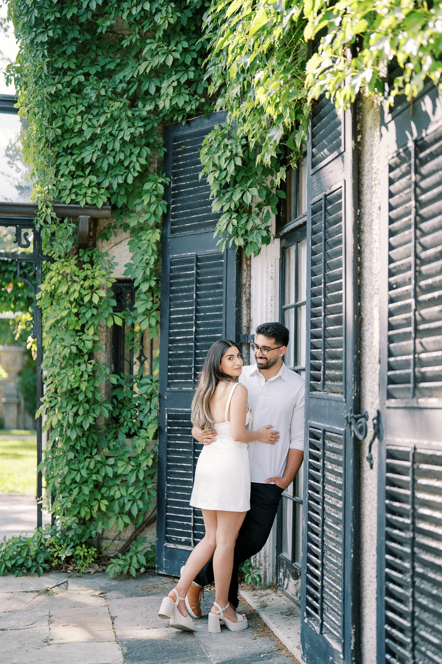 Couple embracing in front of ivy-covered building with black shutters on a sunny day.