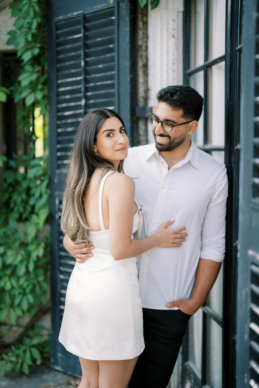 A couple smiling and embracing near a window with greenery in the background. The woman is wearing a white dress and the man is in a white shirt and glasses, creating a romantic and natural setting.