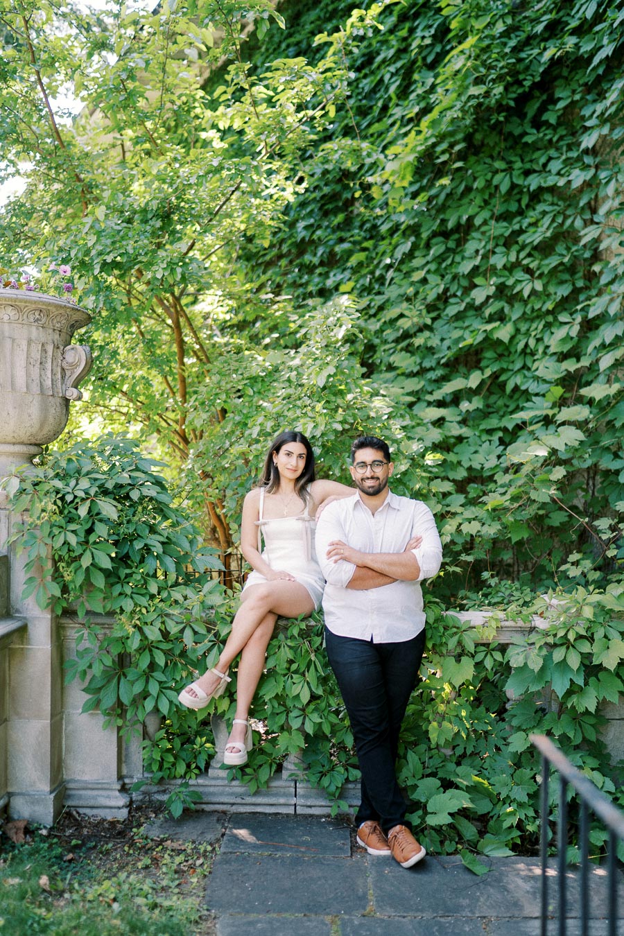 A couple posing in a lush garden setting, surrounded by green foliage and stone architecture, with the woman sitting on a ledge and the man leaning against it.