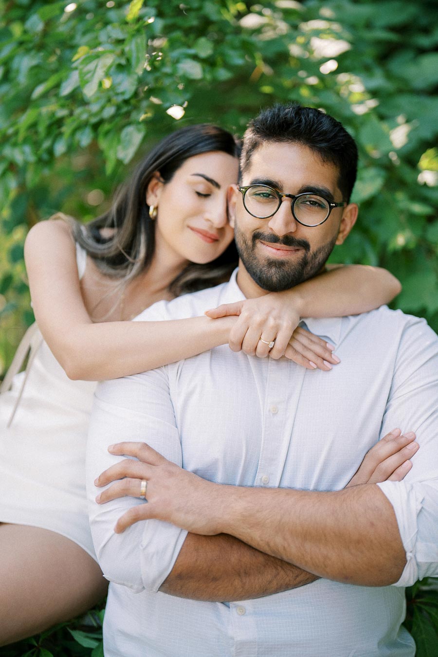 A happy couple embracing outdoors in a lush green setting, with the woman resting her head on the man's shoulder, both smiling contently.