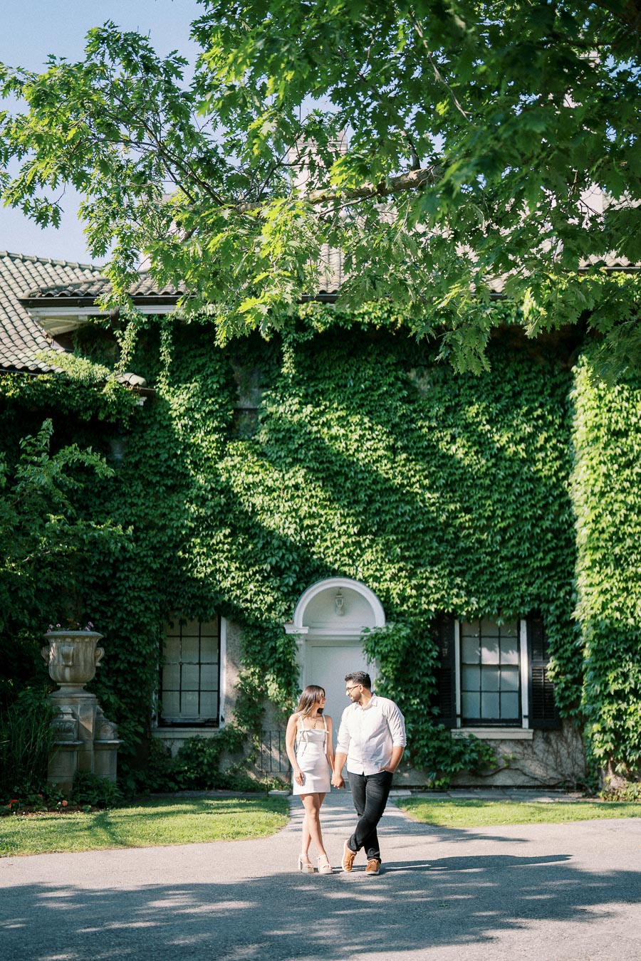 A couple holding hands walking in front of an ivy-covered house with a lush garden on a sunny day.