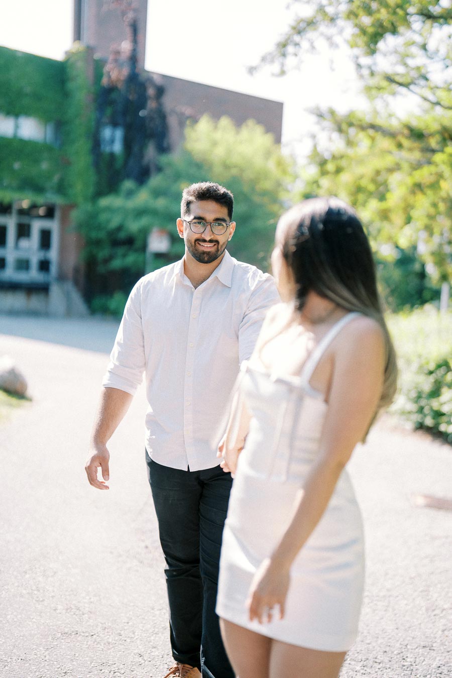 Man and woman walking outdoors on a sunny day, with a blurred background of greenery and a building. Man wearing a white shirt and black pants, while woman wears a light-colored dress.