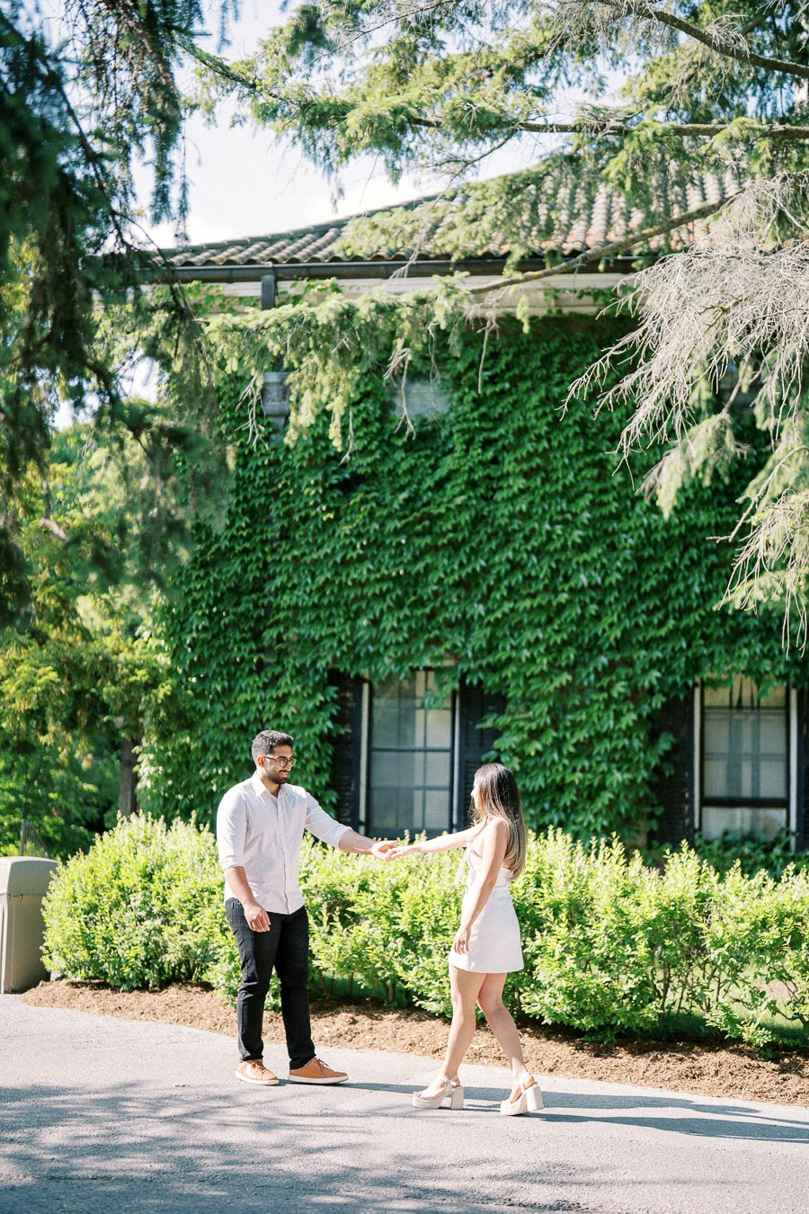 A couple holding hands in front of an ivy-covered building, surrounded by lush greenery and trees on a sunny day.