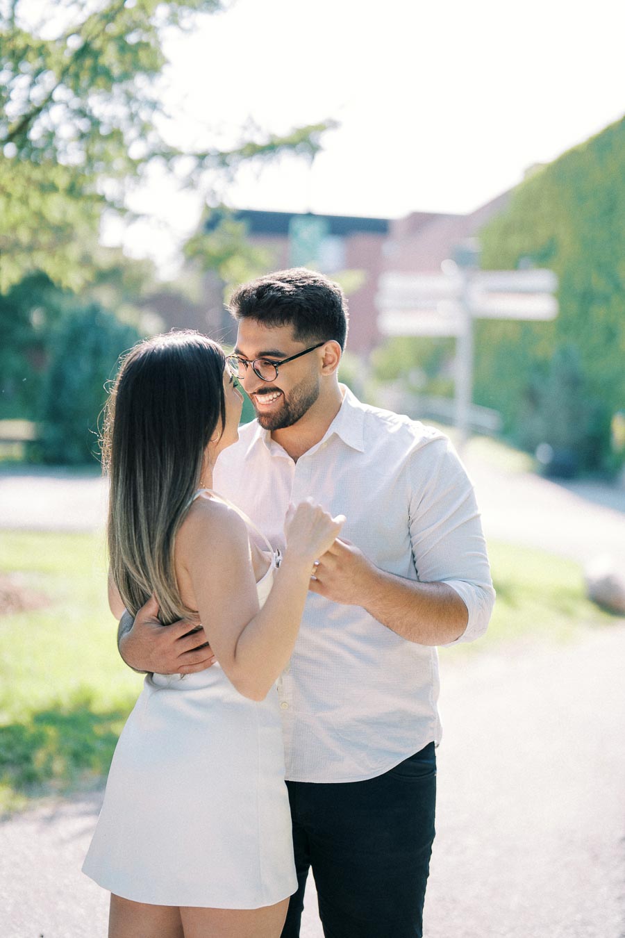 A couple smiling and embracing outdoors on a sunny day, with trees and greenery in the background, capturing a loving moment.