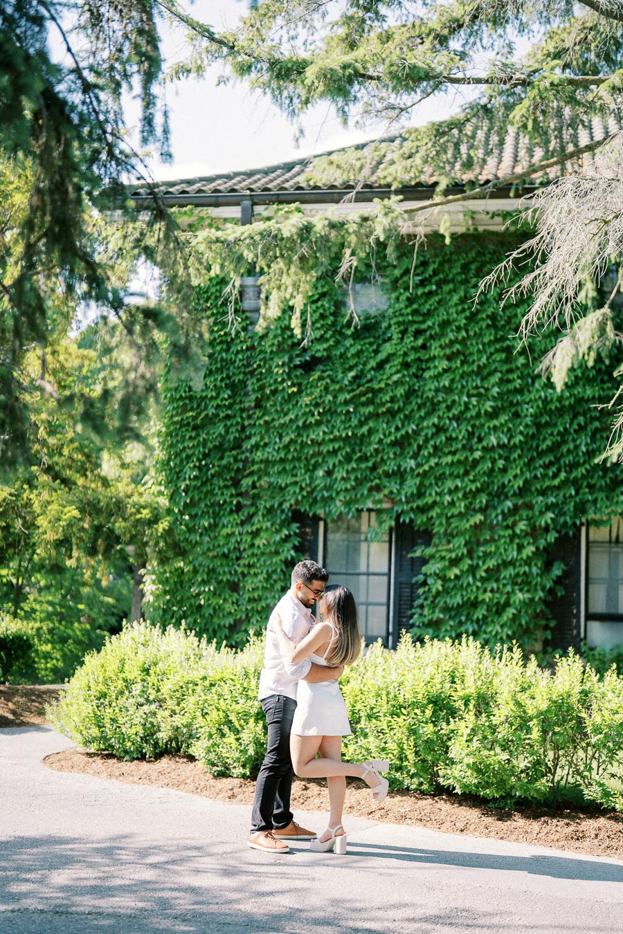 A couple embracing in a sunny garden setting, with a building covered in green ivy in the background and surrounded by lush greenery.
