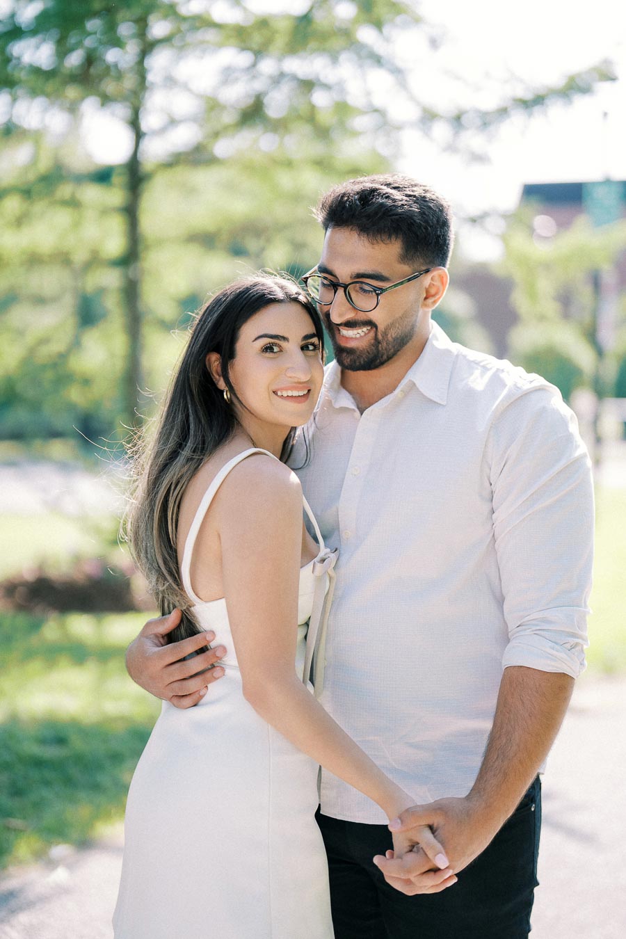 Young couple embracing outdoors in a sunny park, smiling and holding hands, wearing casual white outfits.