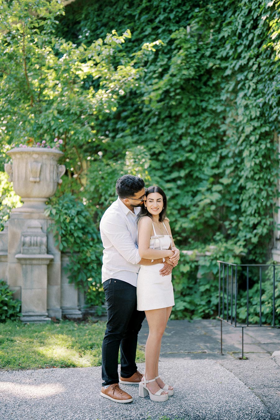 A couple embraces in a sunlit garden, surrounded by lush greenery and a stone planter, creating a romantic and serene atmosphere.