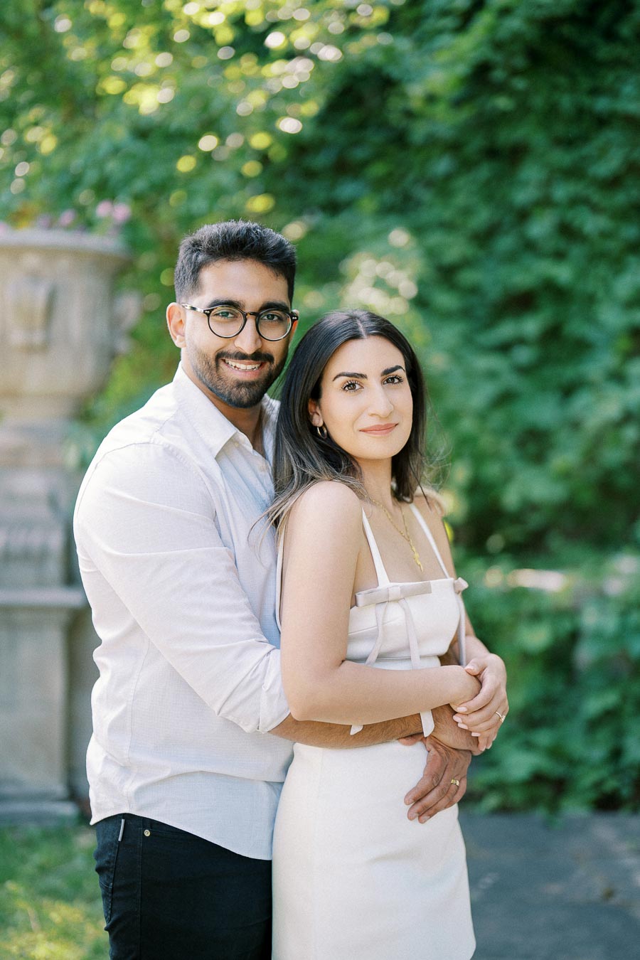 A couple embracing in a garden setting, with lush greenery in the background, both smiling and wearing light-colored summer attire.