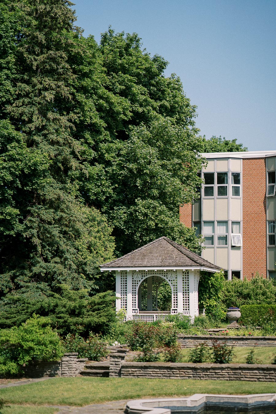 White gazebo surrounded by lush green trees and bushes in a garden setting, with a brick building in the background on a sunny day.